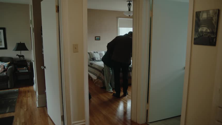 A man helps an elderly woman into the bedroom