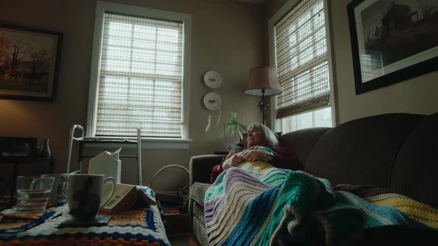 An elderly woman is lying on a sofa with a blanket over her