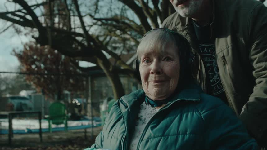 Elderly woman smiles while sitting in a wheelchair pushed down sidewalk leading from house