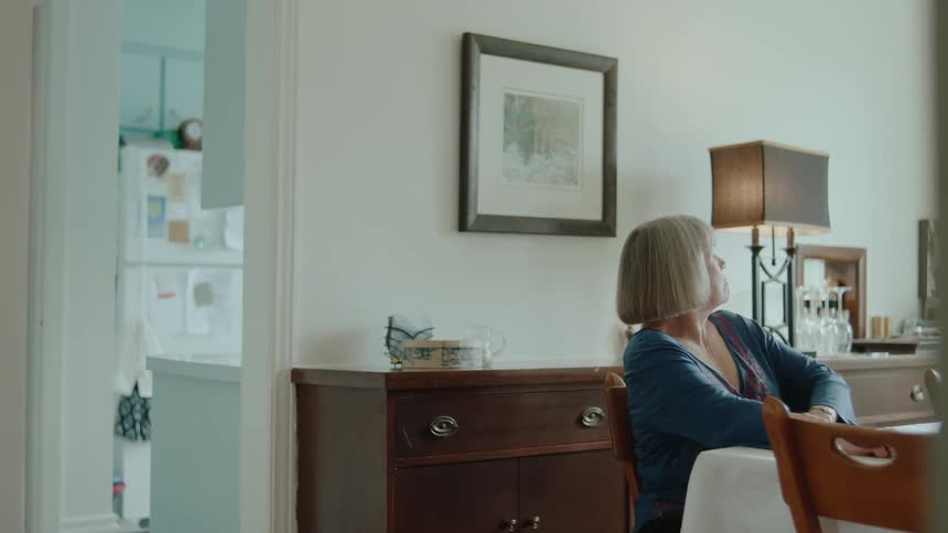 An old woman sits at a dining room table looking sad