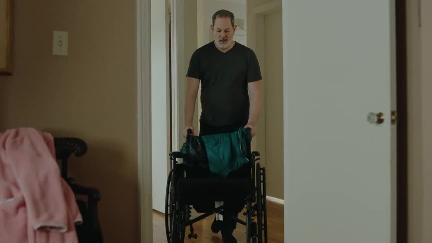 Standing man passing through the hallway and entering a room carrying a wheelchair