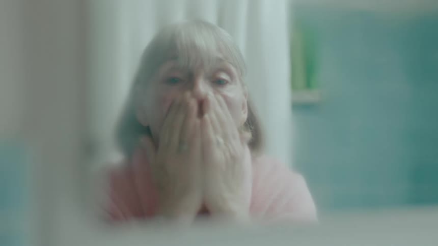 Elderly woman wipes her face while facing herself in the bathroom mirror