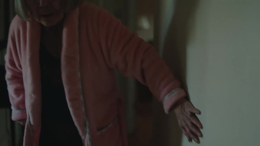 Elderly lady in pink bathrobe walking down the hallway of the house leaning on the walls