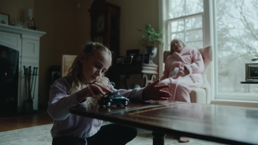 A little blonde girl is playing with a toy car on a table in a living room and behind her is a lady sleeping on the armchair