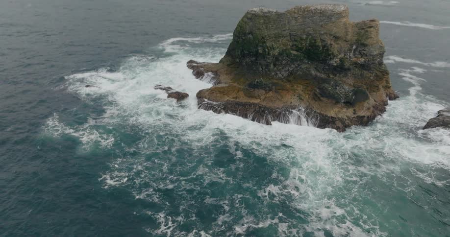 Rocky formations protruding from the ocean with waves crashing against them under a cloudy sky