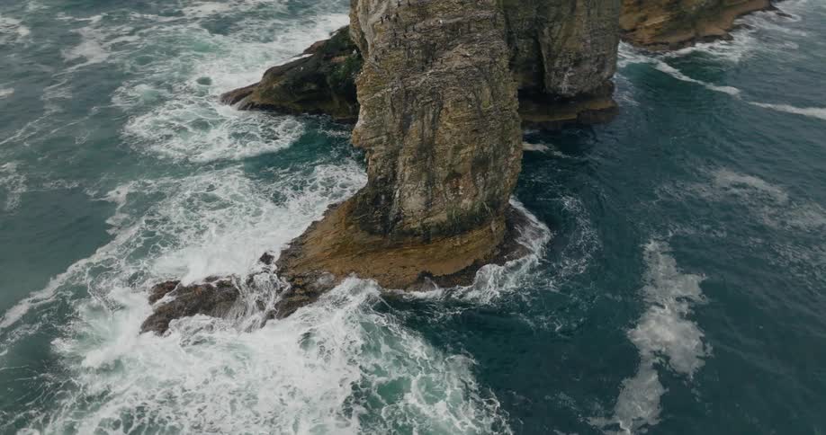 The aerial view shows turquoise waves crashing against a dark rugged rock formation