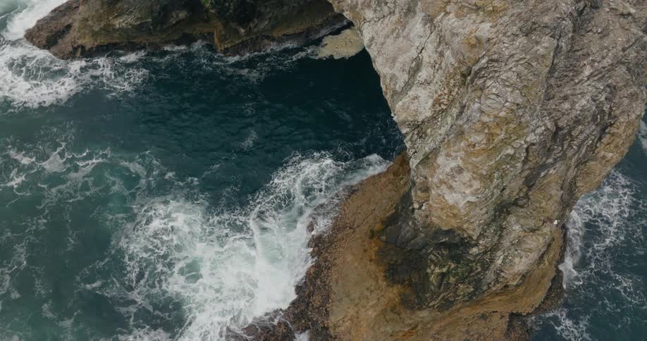 Seagulls fly over a rock arch formation as waves splash against the rock