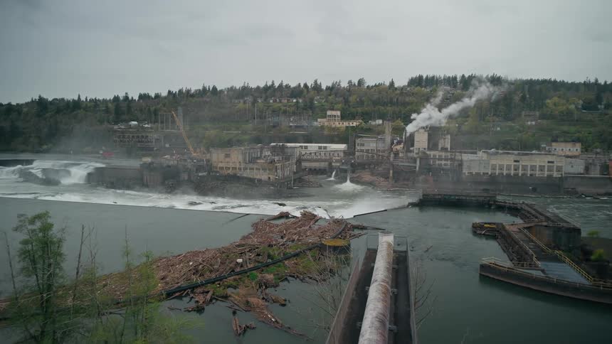 A water treatment unit with a large flow of water passing through an uneven level and behind buildings and one of them is emitting smoke