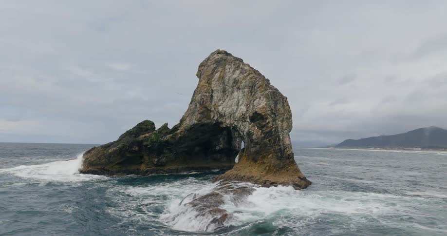 A rocky arch formation stands in turbulent ocean waves under a cloudy sky