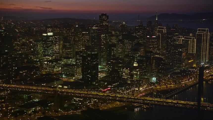 Aerial scene of a bridge with vehicle traffic on the side and a large metropolis with modern buildings illuminated at night and the seafront