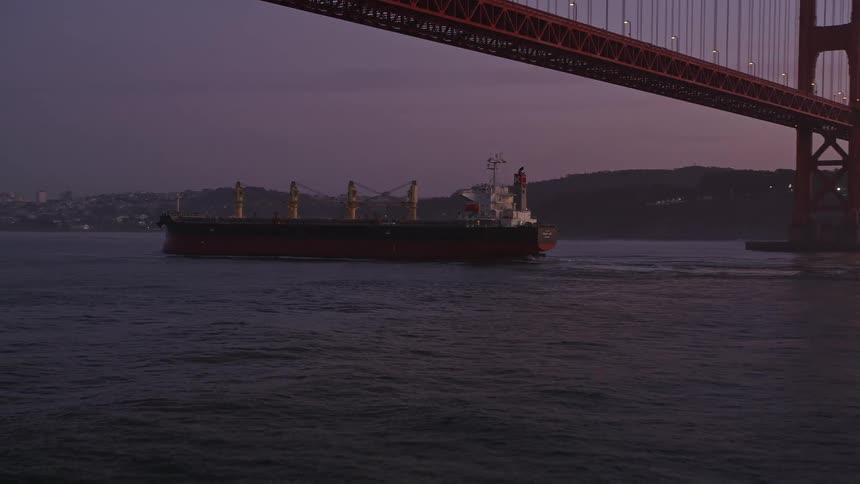 A cargo ship with containers sails near a large bridge during dusk