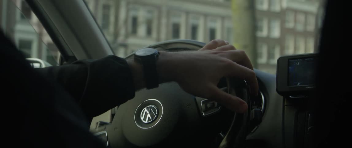 As the man wearing a wristwatch and a black jacket navigates the city streets he firmly grips the steering wheel of his car