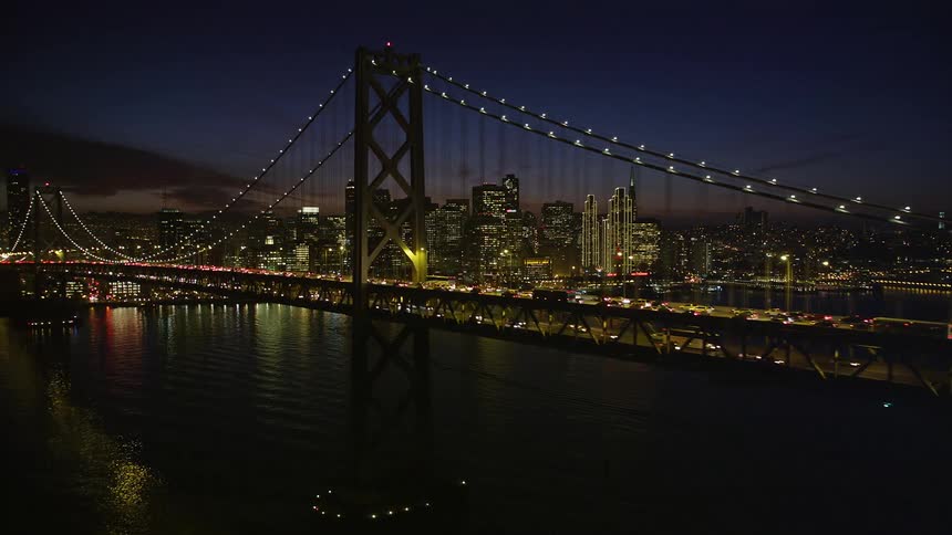 At night an aerial view captures a bridge illuminated with yellow lights while streaks of red and white light trail behind vehicles crossing it