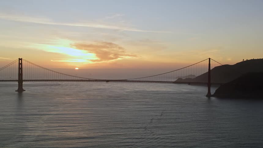 Suspension bridge stretching across the water as the sun glows through clouds in the distance