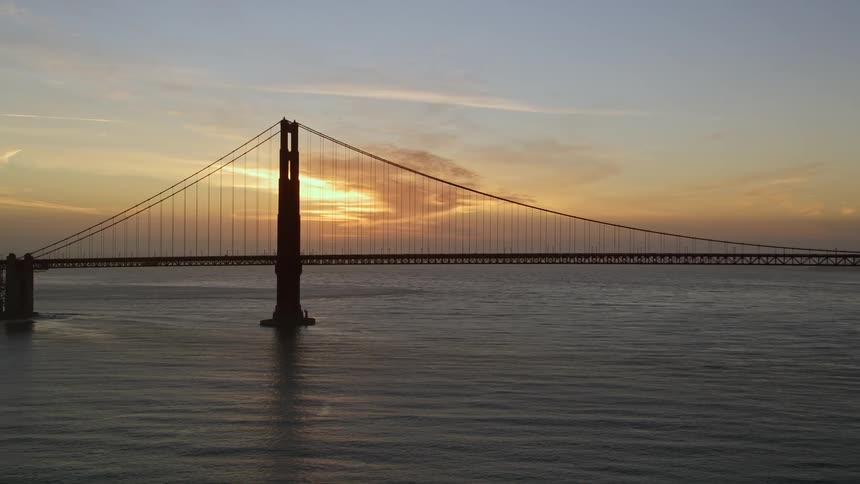 Suspension bridge stretching over the water as the sun glows through clouds in the distance