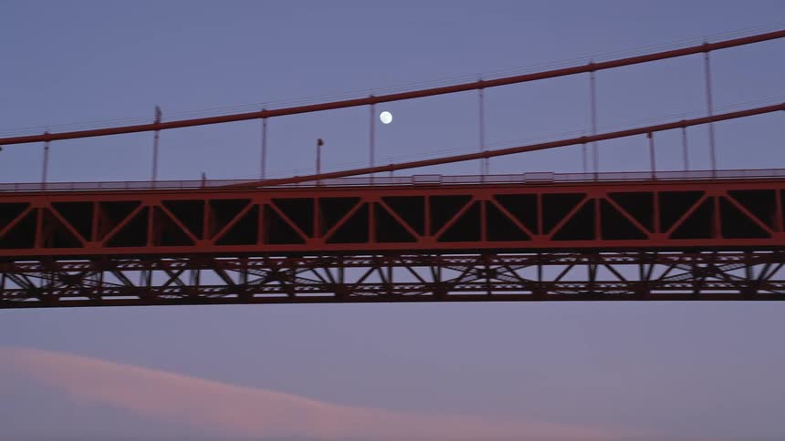 Moon hovers over suspension bridge in the blue sky after dusk