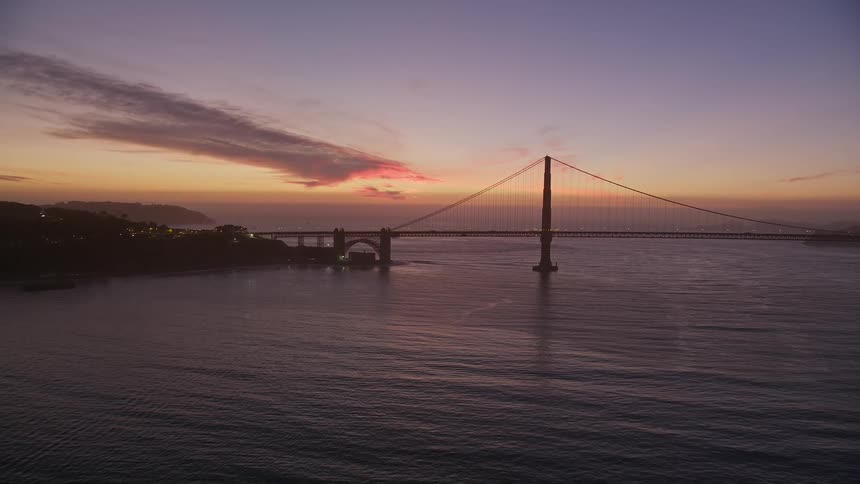 The iconic golden gate bridge gleams orange in the light of the setting sun with wispy clouds above and the calm waters of the bay below