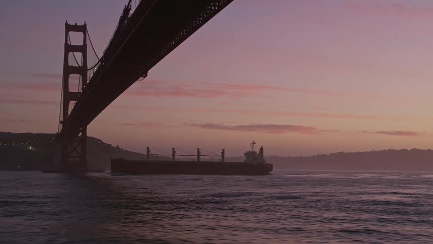 A cargo ship with containers sails near a large bridge during dusk