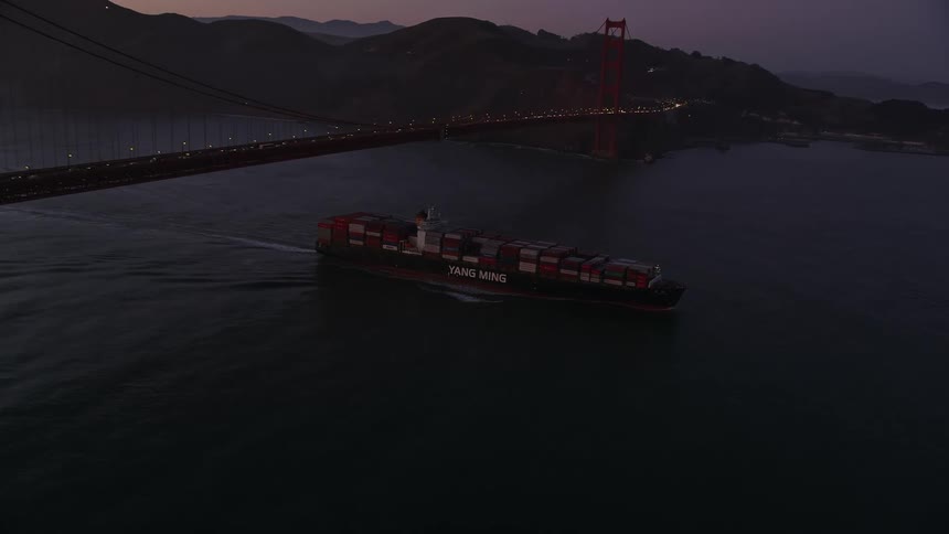 A cargo ship with containers sails near a large bridge during dusk