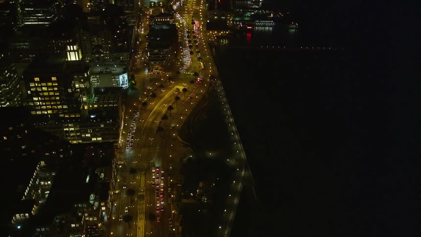 This aerial view shows a network of highways cutting through a vibrant city at night with the empire state building lighting up the midtown skyline