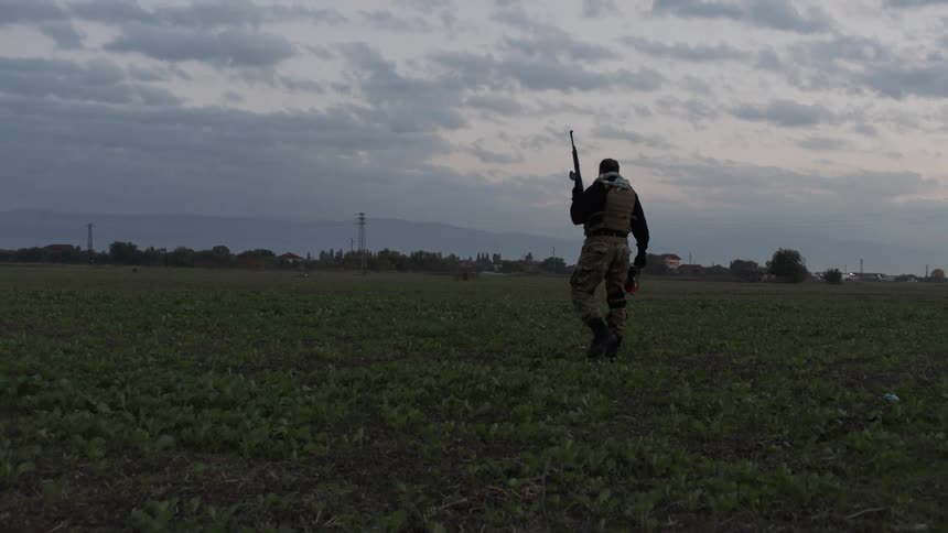 Man with an automatic rifle walks into an empty field at dusk
