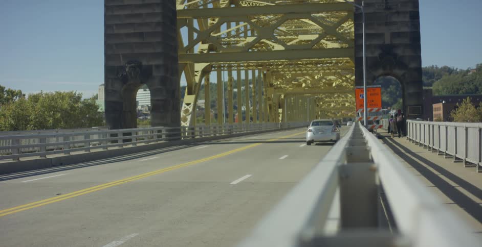 Cars crossing a bridge while people walk down the sidewalk on the edge of the bridge