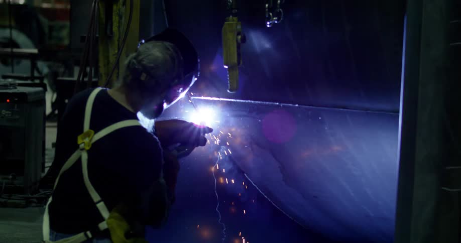 A worker with his back is wearing a helmet while welding a part and a lot of sparks are coming out