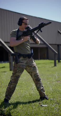A man in camo fires an automatic rifle outside a building