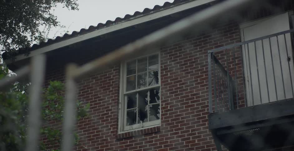 A window in a brick house has broken glass and in front there is an iron gate