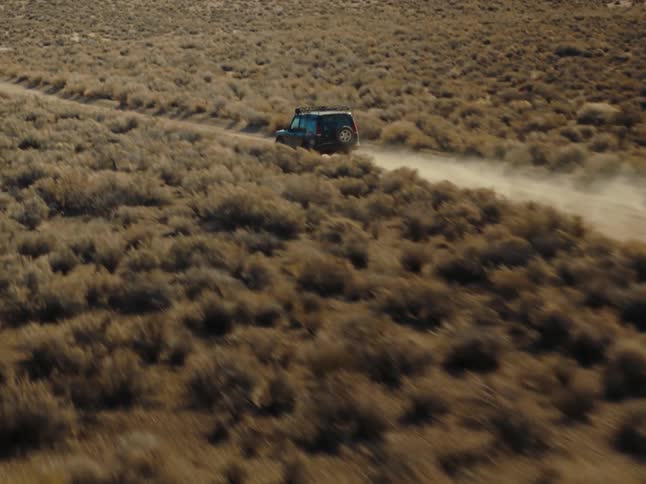A blue jeep is driving through a field of dry grass creating a trail of dust