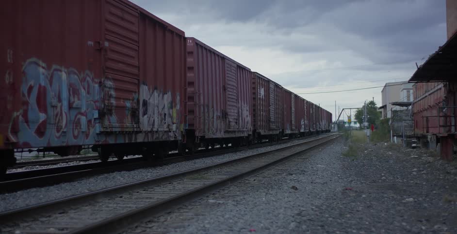 A long train moves down tracks by rundown buildings