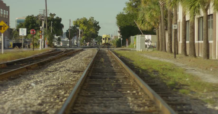 The tracks of a train with a yellow train in the background in an area with some buildings and trees with green leaves