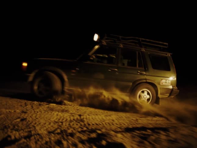 A vehicle with headlights on is driving through a sandy terrain at night