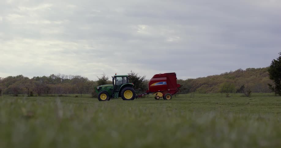 Green tractor pulls trailer across field bordering hill on an overcast day