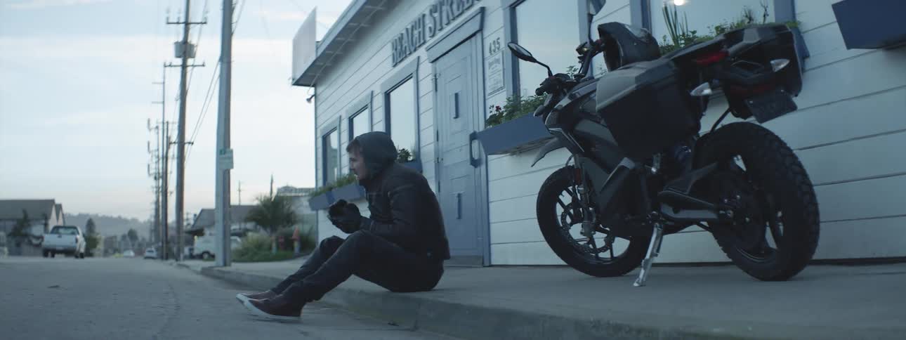 A man sits on a sidewalk next to a motorcycle and blows on his hands to keep warm