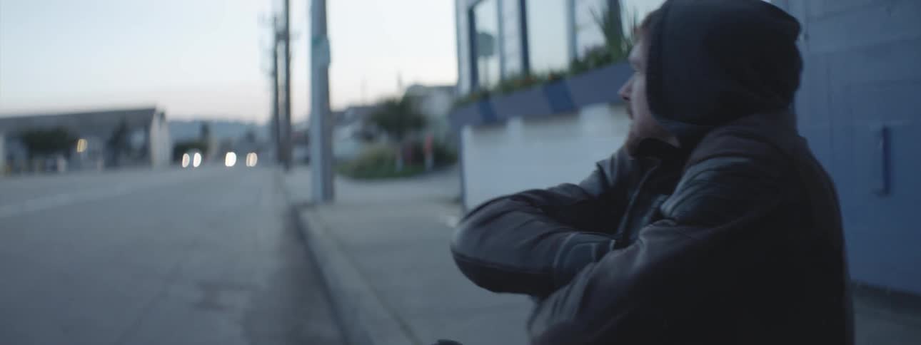 Man in a leather jacket and gloves sitting cold on the sidewalk of a street