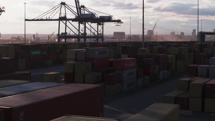 Several containers stacked in a port with some huge cranes in the background