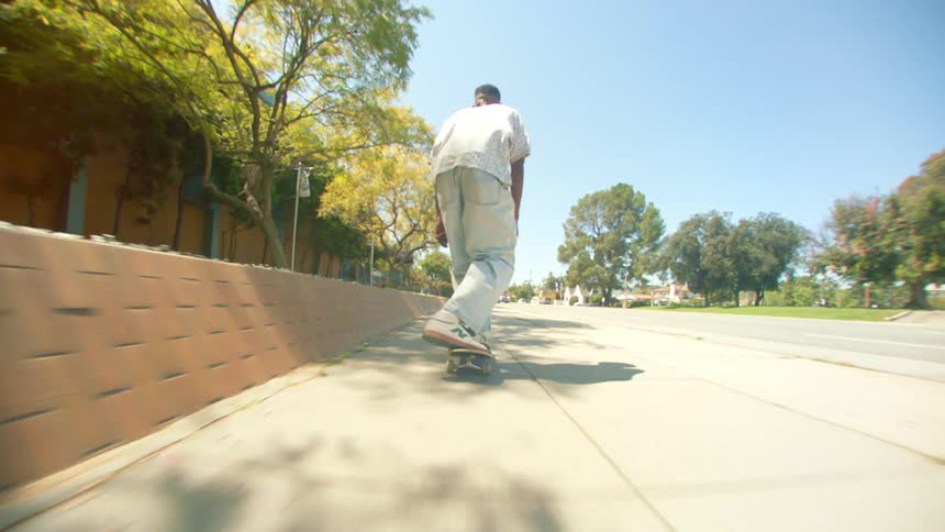 A man from behind is wearing a white shirt and is riding a skateboard on the street and then does a gauntlet