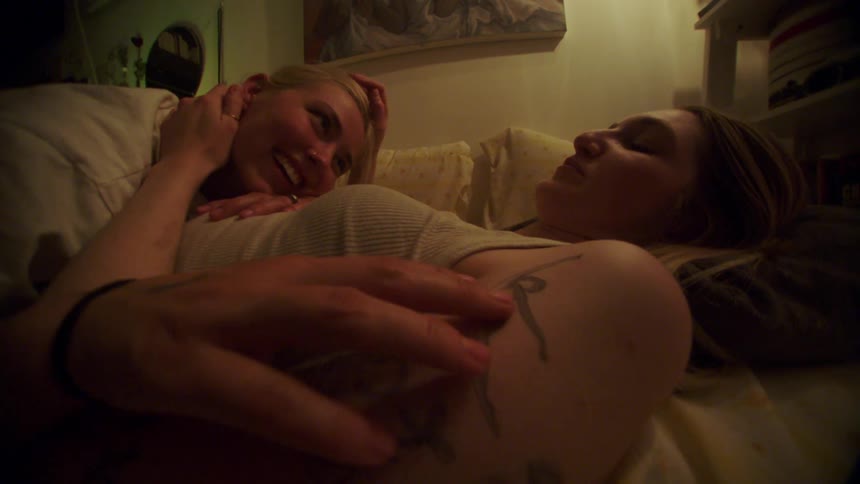 Two young women lying in bed together caressing each other while smiling and talking