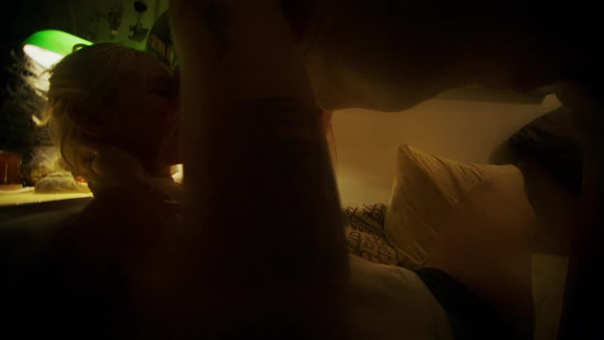A blonde woman wearing a white shirt and black shorts makes out with another woman who is wearing a white top and shorts in the bedroom