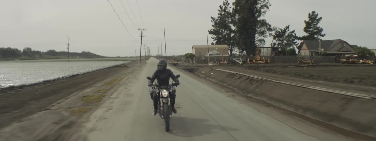 A person in dark attire and a helmet rides a motorcycle down a narrow road flanked by fields