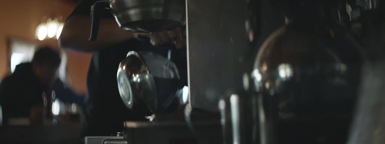 A barista is preparing coffee by holding a pot under a coffee machine in a dimly lit setting