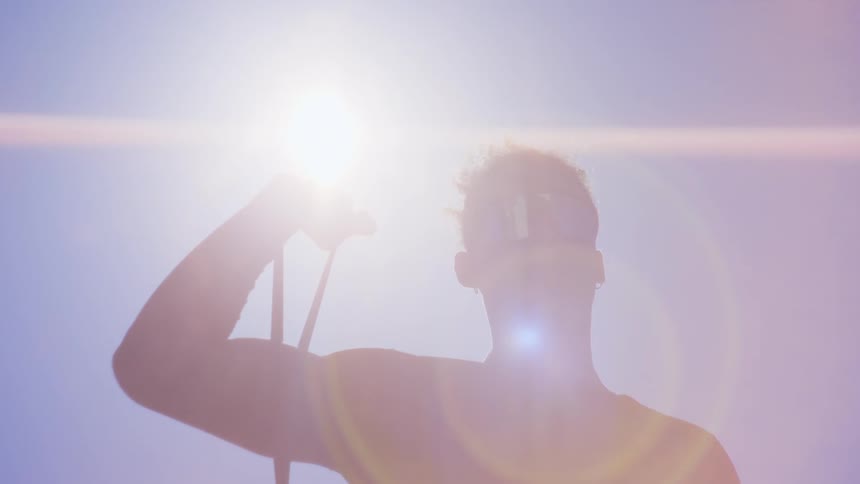 A young man wearing reflective glasses is in a swimsuit holding a rope and in contrast with the sunlight in the sky