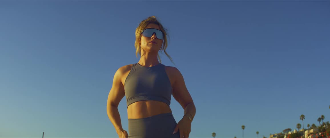 Blonde woman wearing sunglasses sports bra and leggings stands against the blue sky