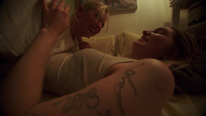 A blonde woman wearing a white shirt makes out with another woman lying down on the bed laughing together