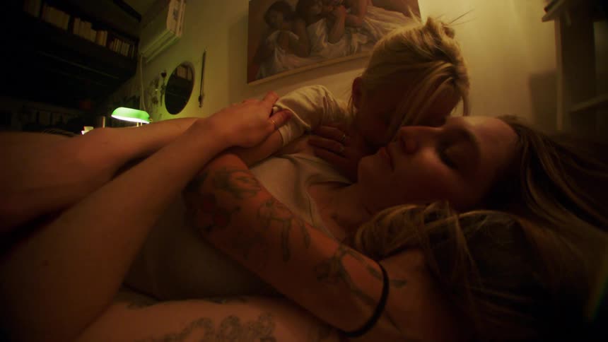 A blonde woman with a white tshirt and a tattoo holds and kisses a woman lying on a bed in the bedroom