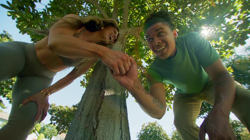 A woman in a sports bra and leggings and a man in a green tshirt smile and hold hands under the canopy of a sunlit tree creating a joyful and connected moment