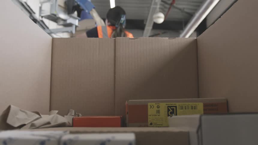 A worker in an orange vest is packing a box with various items