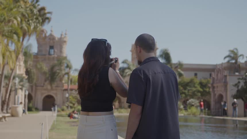 Woman takes a picture of landmark buildings while standing near a pond with a man