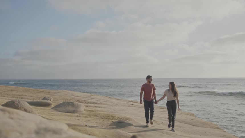 A bearded man wearing a red tshirt and dark blue pants walks hand in hand with a woman in a white shirt and dark blue pants along the beach near the sea